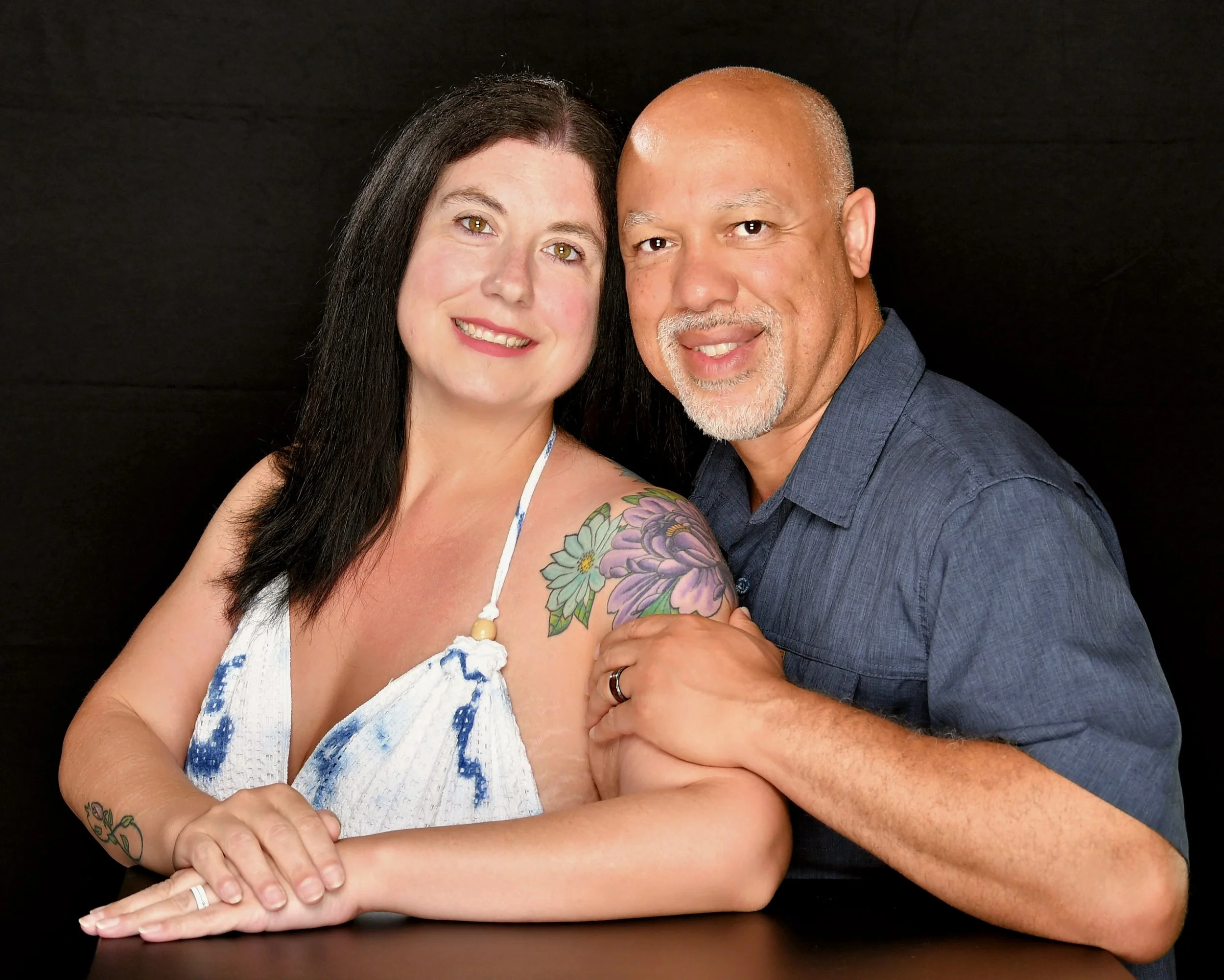 A smiling couple with dark hair and light skin, the woman has tattoos on her shoulder and arm, and is wearing a white and blue dress. The man has a beard and is wearing a blue shirt, holding the woman's hand with a wedding ring visible.