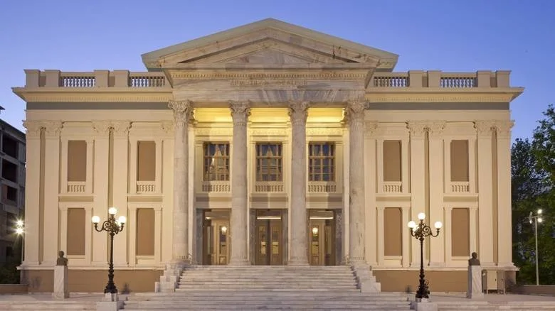 A grand neoclassical building with tall columns, a triangular pediment, and steps leading up to the entrance, illuminated at dusk.