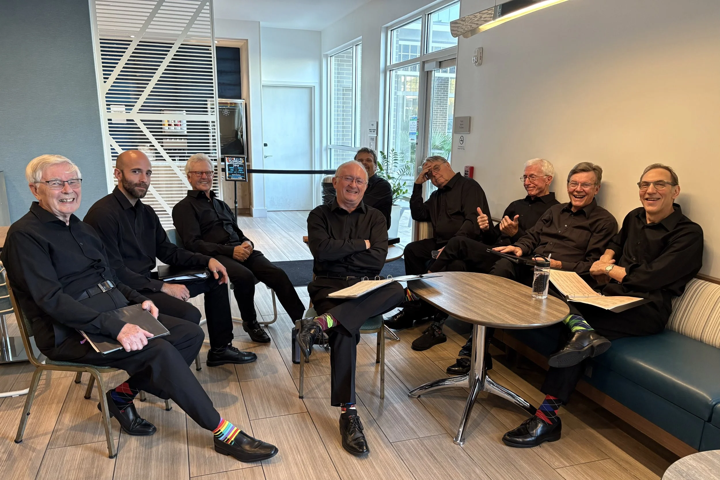 A group of nine men in black shirts and black pants sitting around a small wooden table in a modern room with large windows, some smiling and laughing, with some holding notebooks and pens.