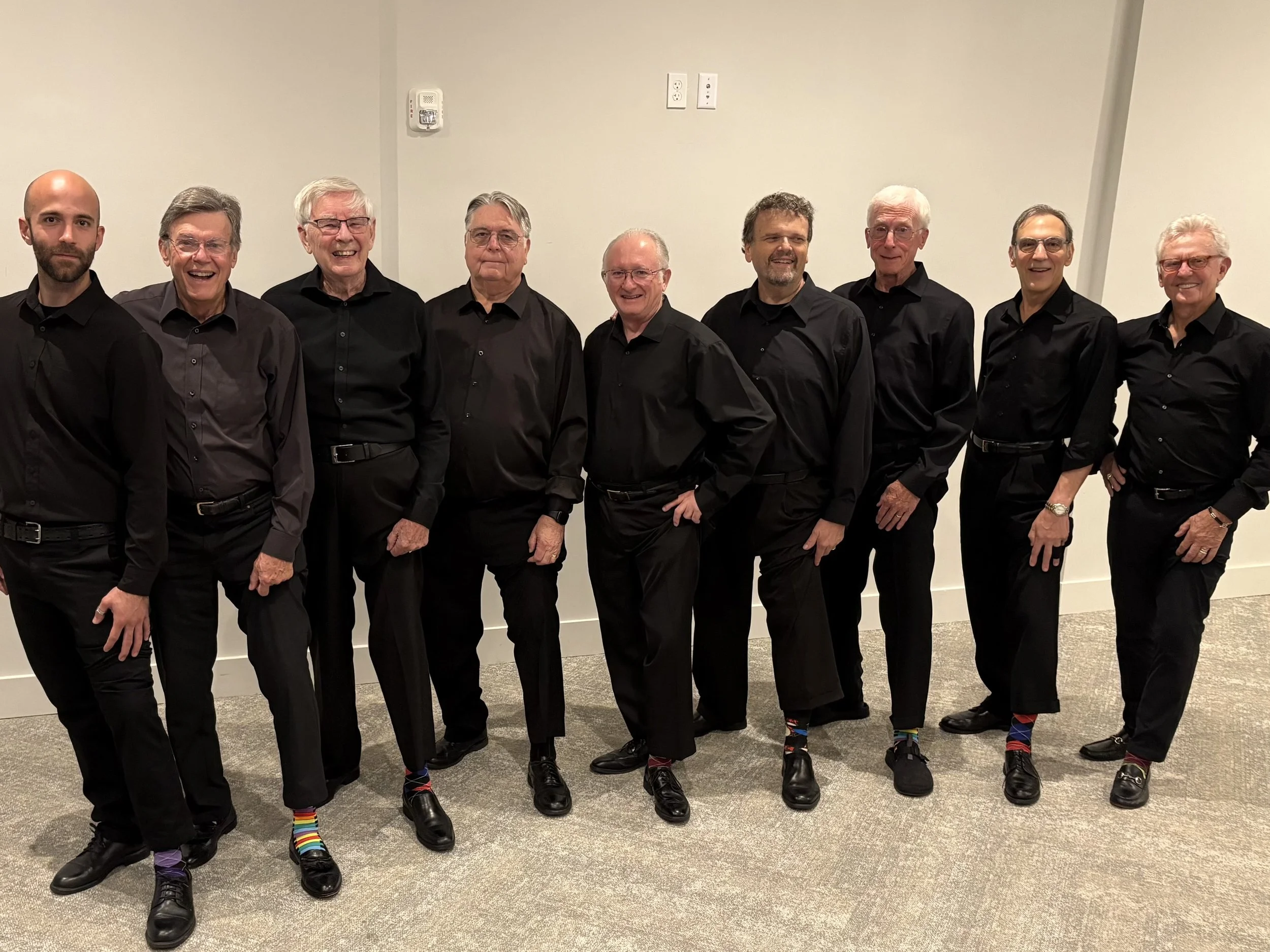 Ten men dressed in black shirts and black pants, standing side-by-side against a plain white wall with electrical outlets, smiling and posing for a group photo.