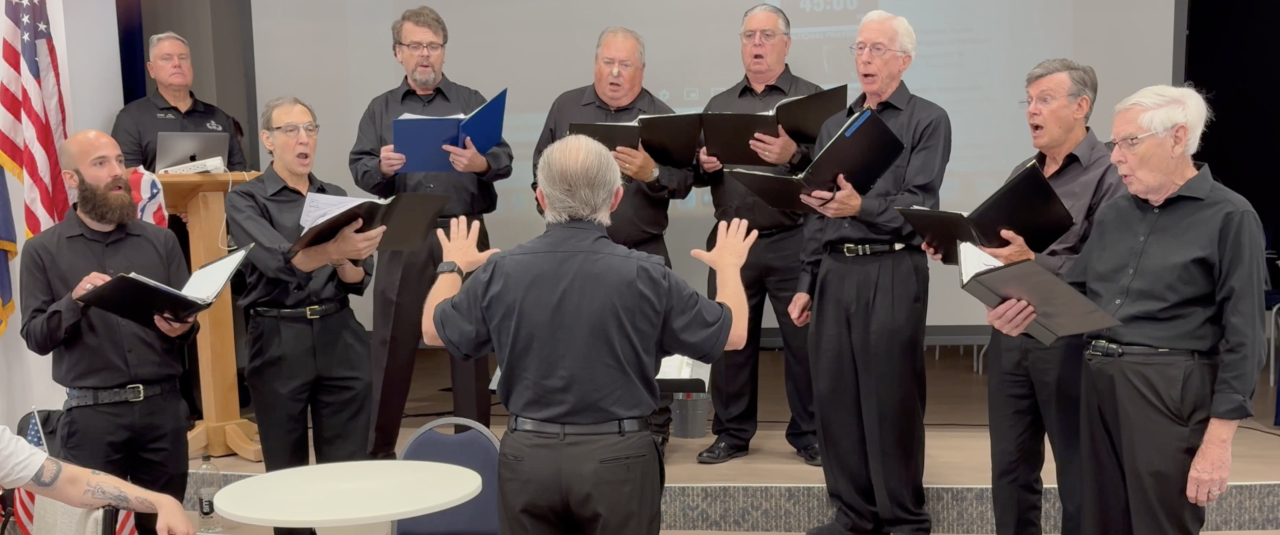 A men's choir performing under the direction of a conductor in a room, with the choir members holding black folders and singing, some with open mouths, in front of a whiteboard and American flags.