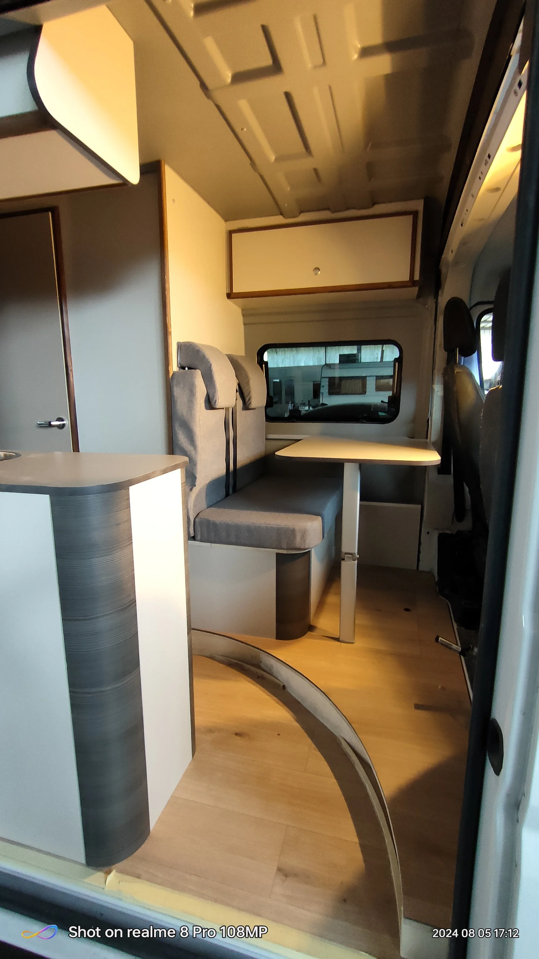 Interno di un camper con zona soggiorno, tavolo e sedili, finestre e armadiature.