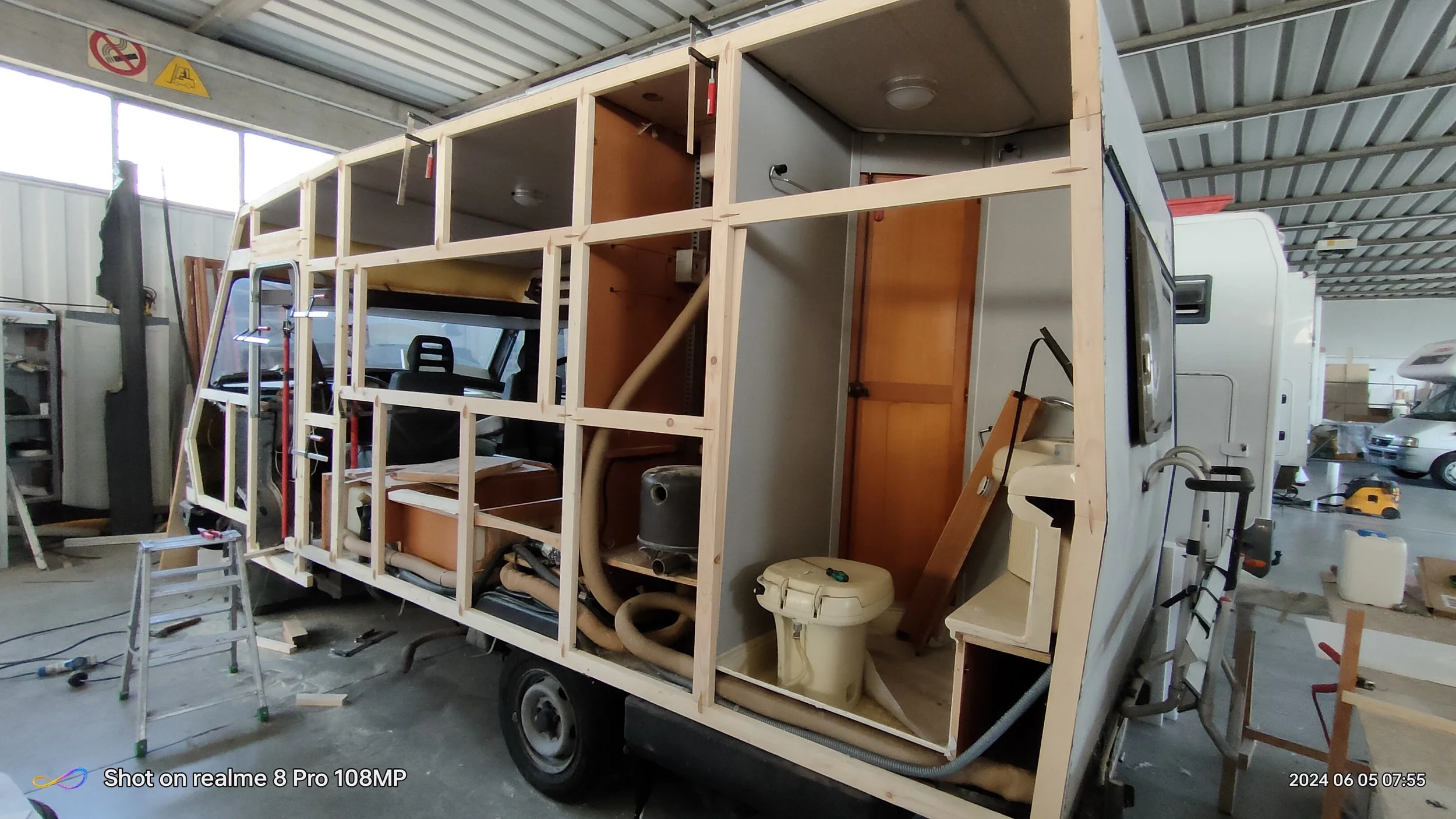 Interno di un'insolita casa mobile in costruzione con struttura in legno visibile, con cucinino e toilette installate, in un capannone di lavoro.