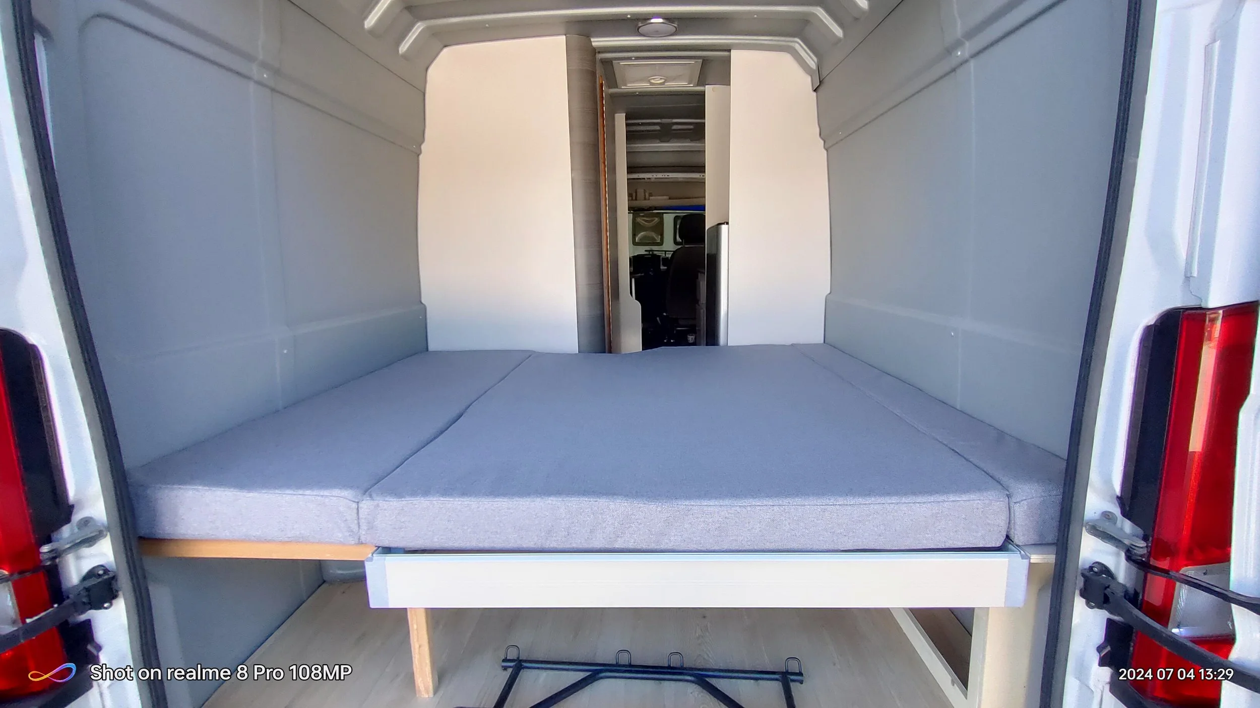Interno di un veicolo modificato, con un'area letto con materasso grigio e un sedile di guida visibile in fondo.