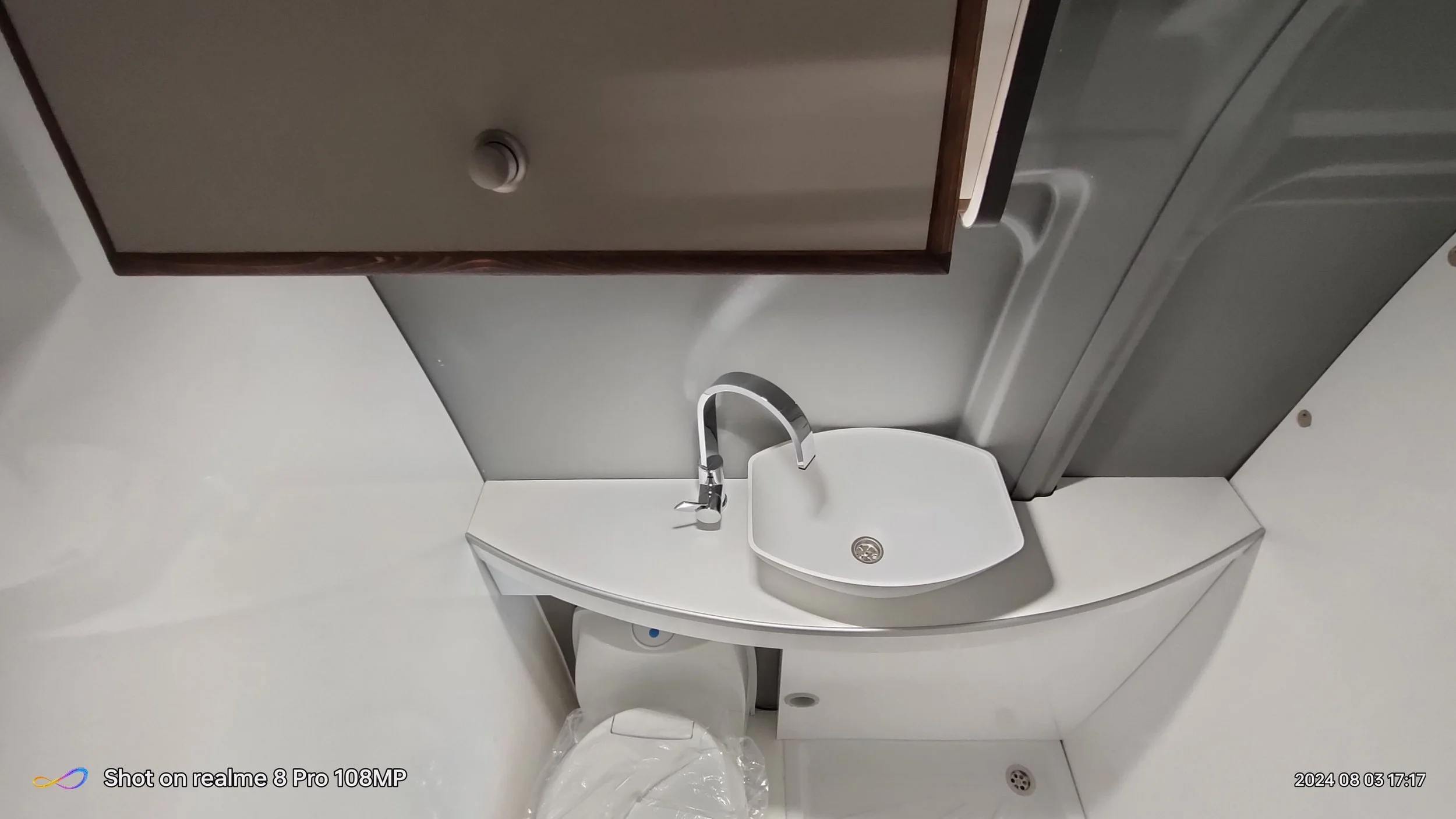 Un piccolo bagno con lavabo bianco a forma di piccola barca, sgorgo con griglia e rubinetto moderno, specchio con cornice in legno chiaro, porta doccia in vetro, water vicino al lavandino, tutto in uno spazio compatto.