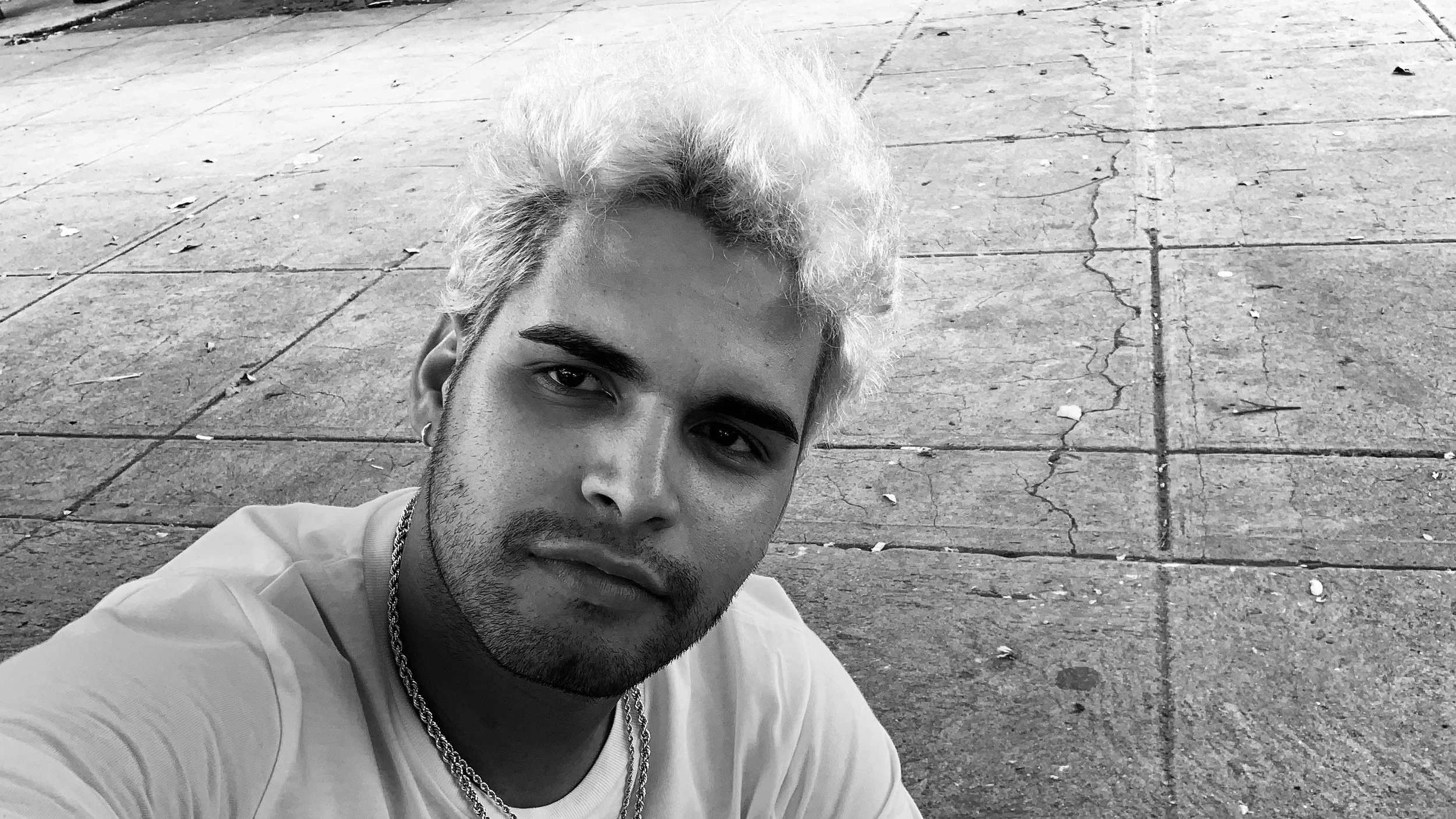 A young man with light-colored hair and a beard, wearing a white shirt and layered necklaces, looks into the camera while sitting on a cracked concrete sidewalk.