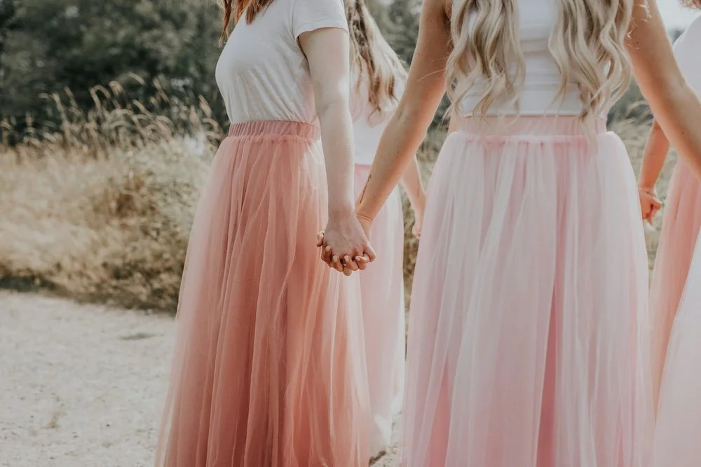 Two women holding hands, wearing white tops and long, flowing pink skirts, outdoors in a natural setting.