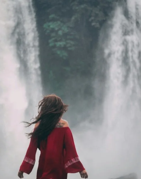 A woman with long hair in a red sweater standing near a waterfall with mist.
