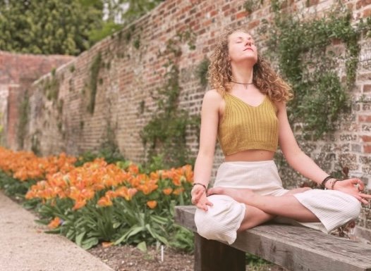 A young woman practicing yoga outdoors, sitting cross-legged on a wooden bench with eyes closed, in front of a brick wall and orange flowers.