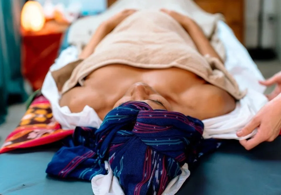 Preparation for womb hara massage treatment.A woman lying on a treatment couch with a beige blanket covering her body. Her head is wrapped in a colorful cloth, and her arms are resting in front of her body.