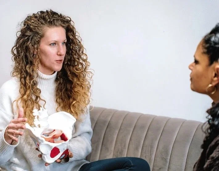 Two women having a serious conversation on a beige couch; one woman with curly blonde hair holding a broken white teddy bear with red spots, wearing a white sweater, and the other woman with dark curly hair listening.
