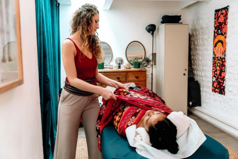 A woman appears to be a massage therapist or healer, holding a bag placed on a reclining woman who is lying on a massage table with a towel over her head in a cozy room with colorful artwork, a mirror, and a cabinet.