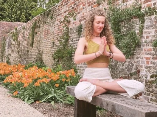 Young woman meditating outdoors on a park bench near orange flowers and a brick wall