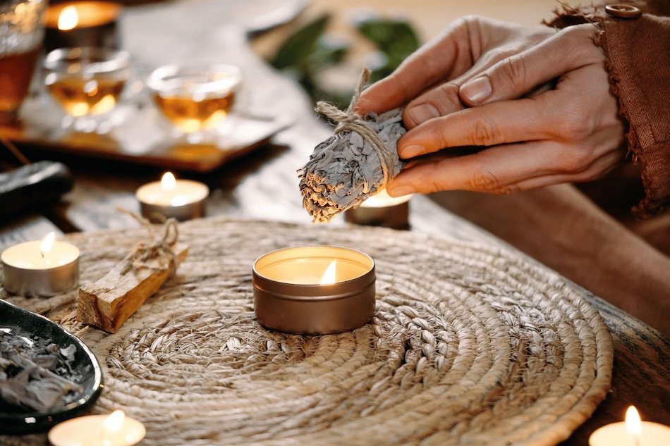 Close-up of a person using a sage bundle to light candles on a woven placemat, with lit candles and drinks in the background.