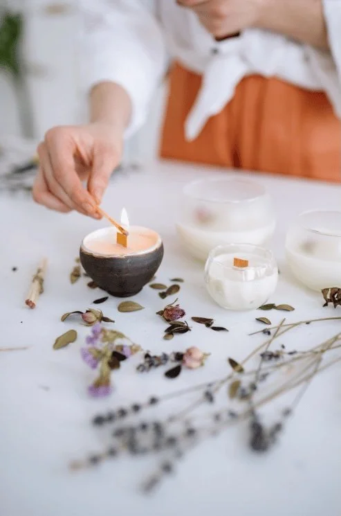 Person lighting a candle in a black bowl surrounded by dried flowers and candles on a white surface.