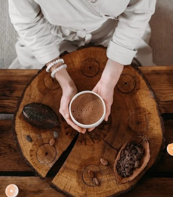 Person holding a cup of cacao over a round wooden table decorated with natural items like a stone, nut shells, and pine cones.
