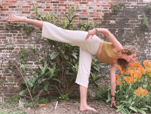 A woman performing a balancing yoga pose outdoors in a garden with plants and orange flowers, against a brick wall.