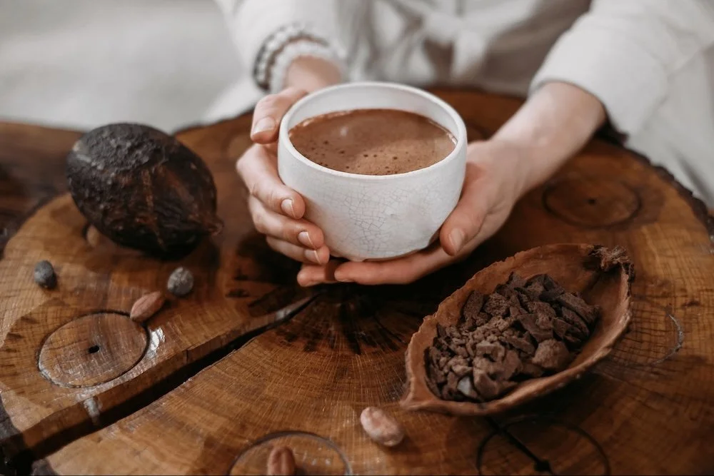 Person holding a white ceramic cup of ceremonial cacao with cocoa powder on top, placed on a rustic wooden table alongside a piece of dark chocolate and a small chocolate-encased item. The table has a natural, textured surface.