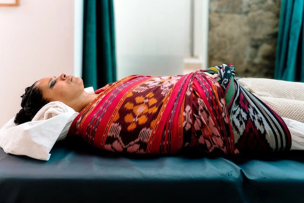 A woman lying on a bed wrapped in a colorful traditional woven blanket during the rebozo womb wrapping also knows as feminine soul retrieval ritual.