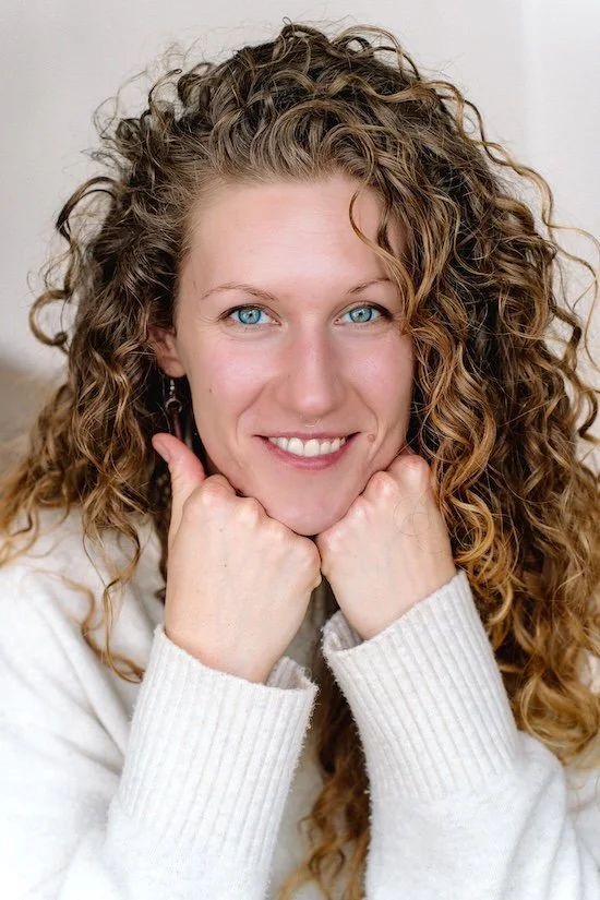 A woman with curly light brown hair and blue eyes smiling with fists under her chin, wearing a white sweater.