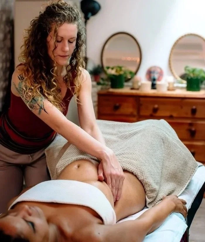 A woman giving an abdominal massage to a person lying on a treatment table in a cozy room with wooden furniture and decorative mirrors.