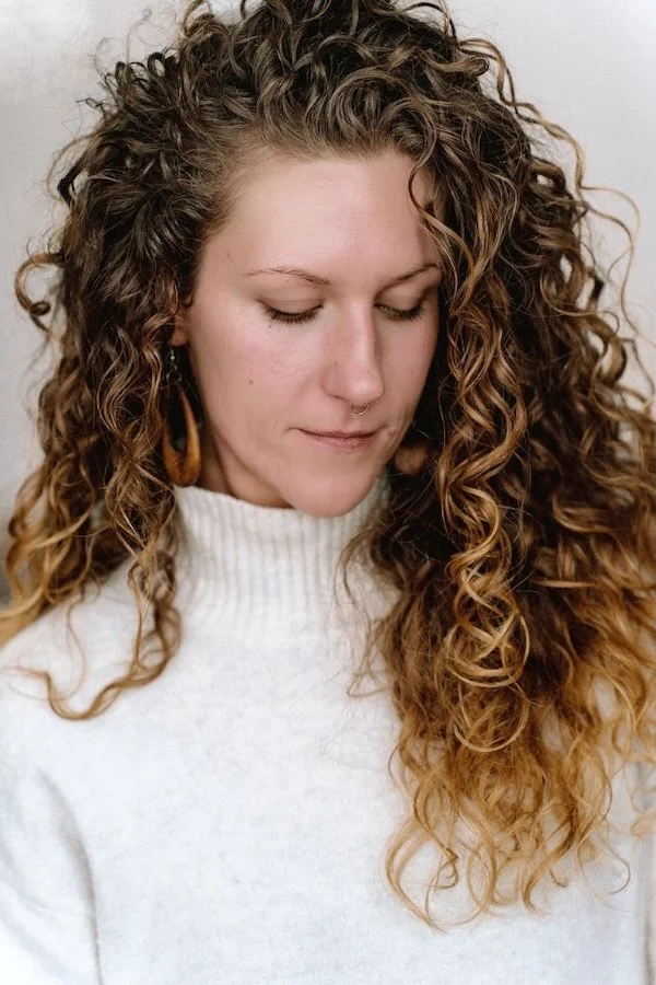 A woman with long, curly brown hair and fair skin, wearing a white turtleneck sweater and earrings, looking downward.