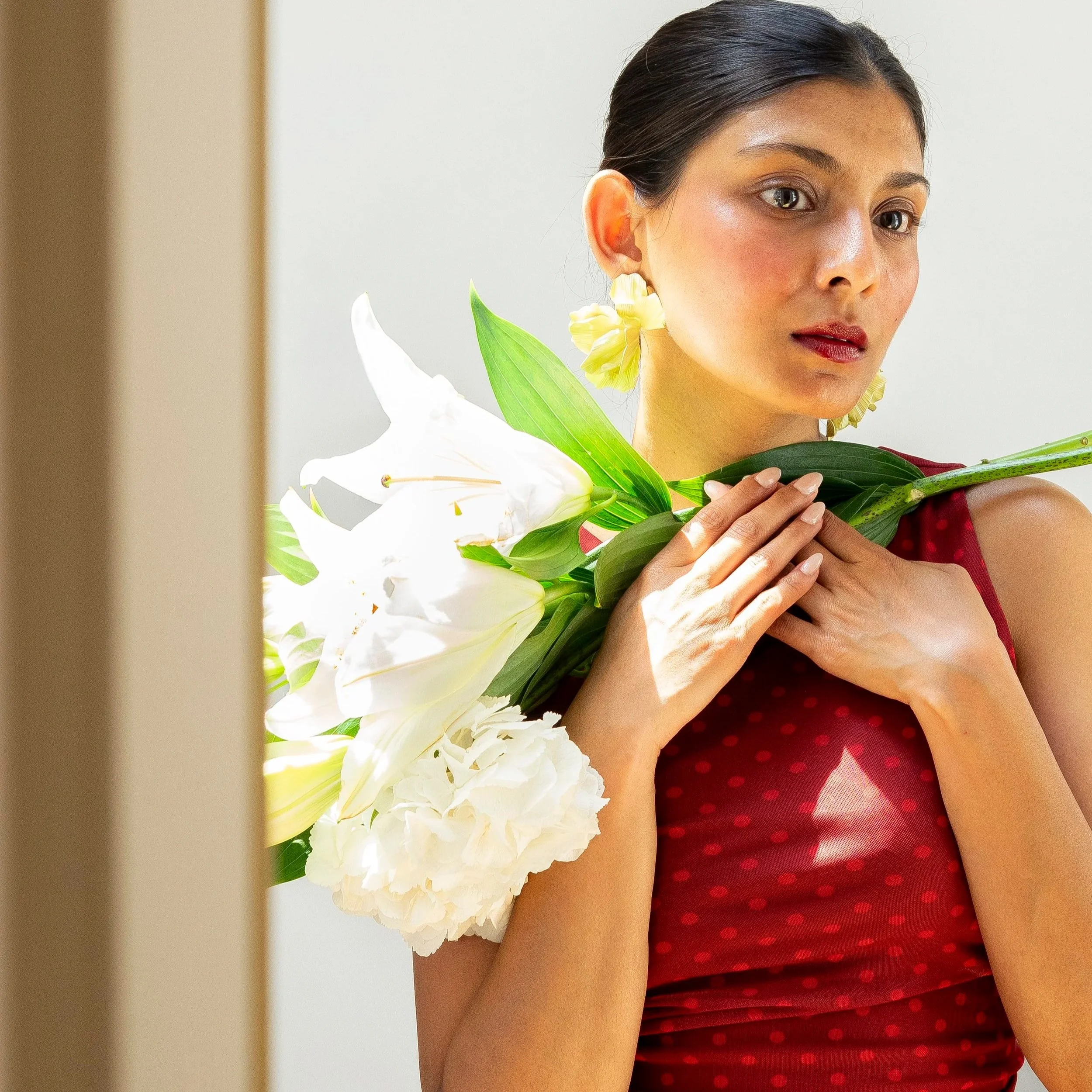 Close-up of a woman's face and upper body, holding white flowers with green leaves, wearing a red polka-dot dress, with dark hair pulled back, in a light-colored background.