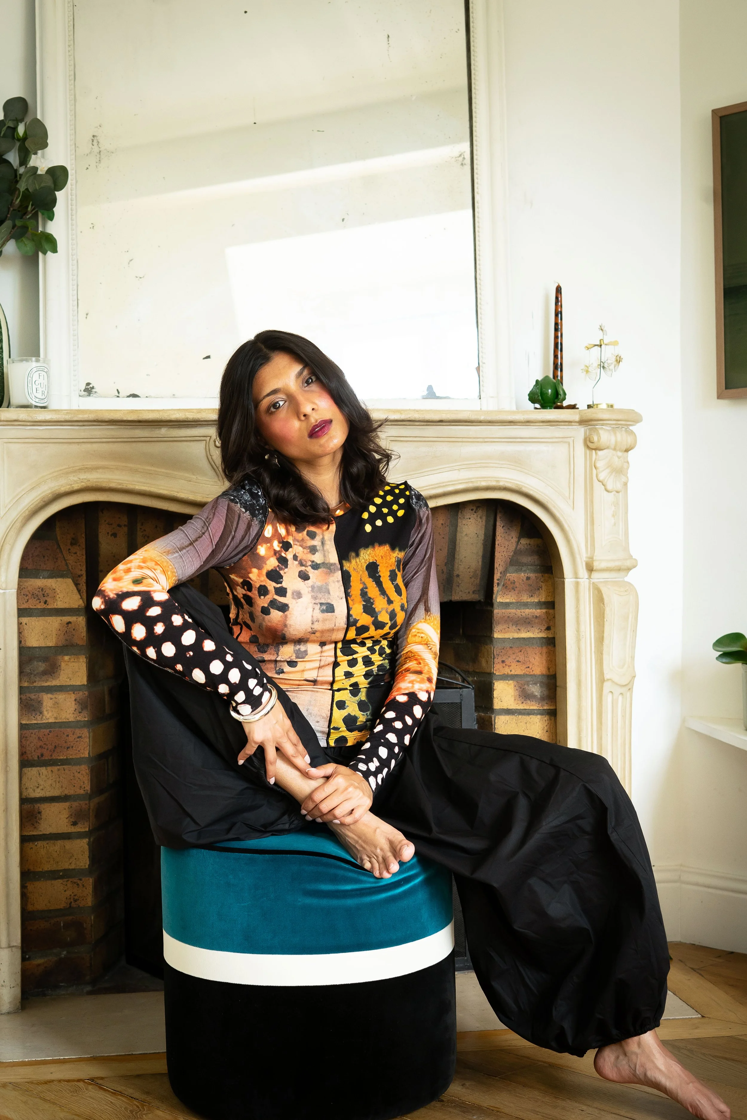 A woman sitting on a colorful stool in front of a fireplace, wearing a multicolored dress with animal print patterns, with black wide-leg pants, barefoot, with her arm resting on her knee, and looking at the camera.