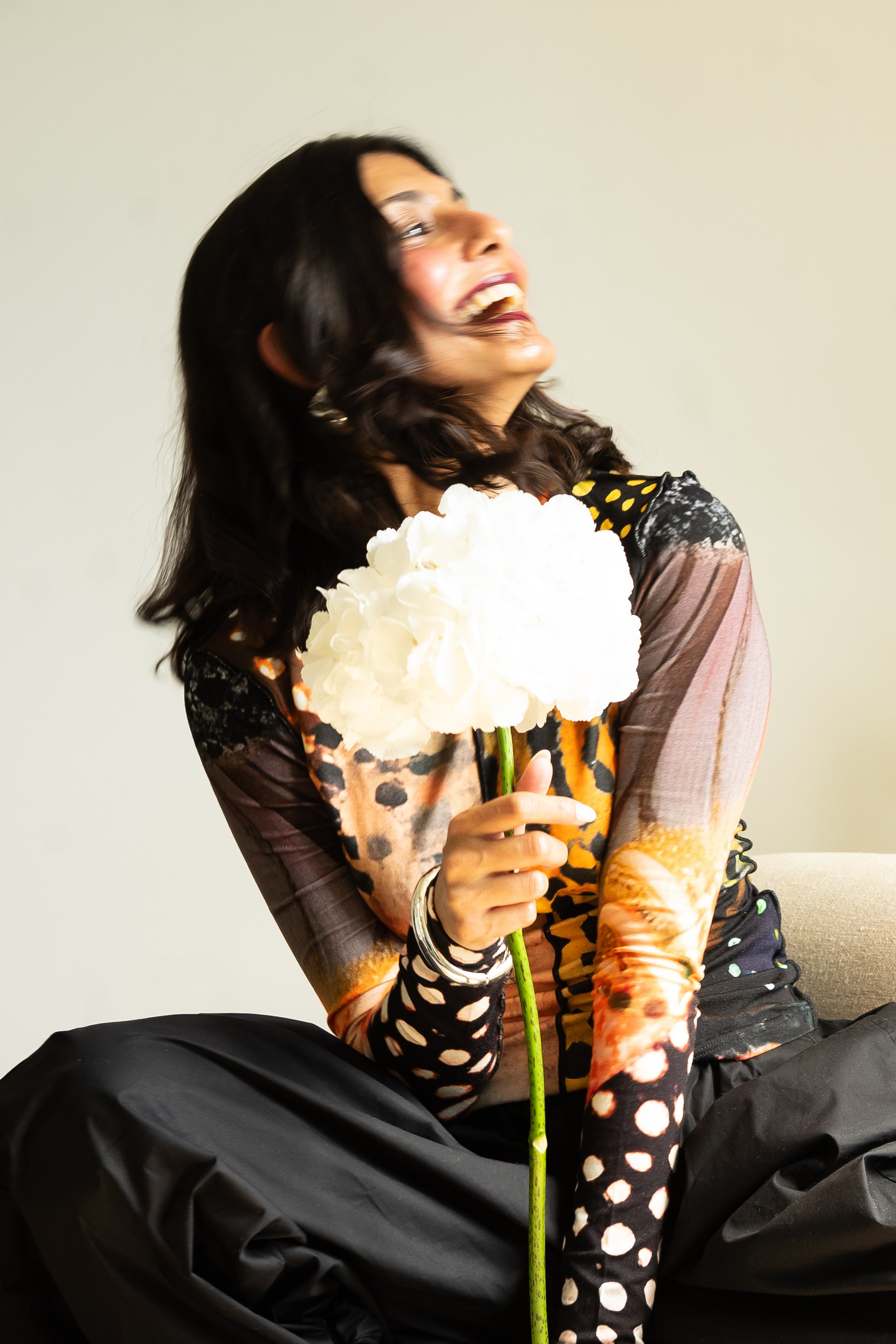 A woman with dark hair, wearing colorful patterned clothing, sits on a beige couch holding a large white fluffy flower and laughs.