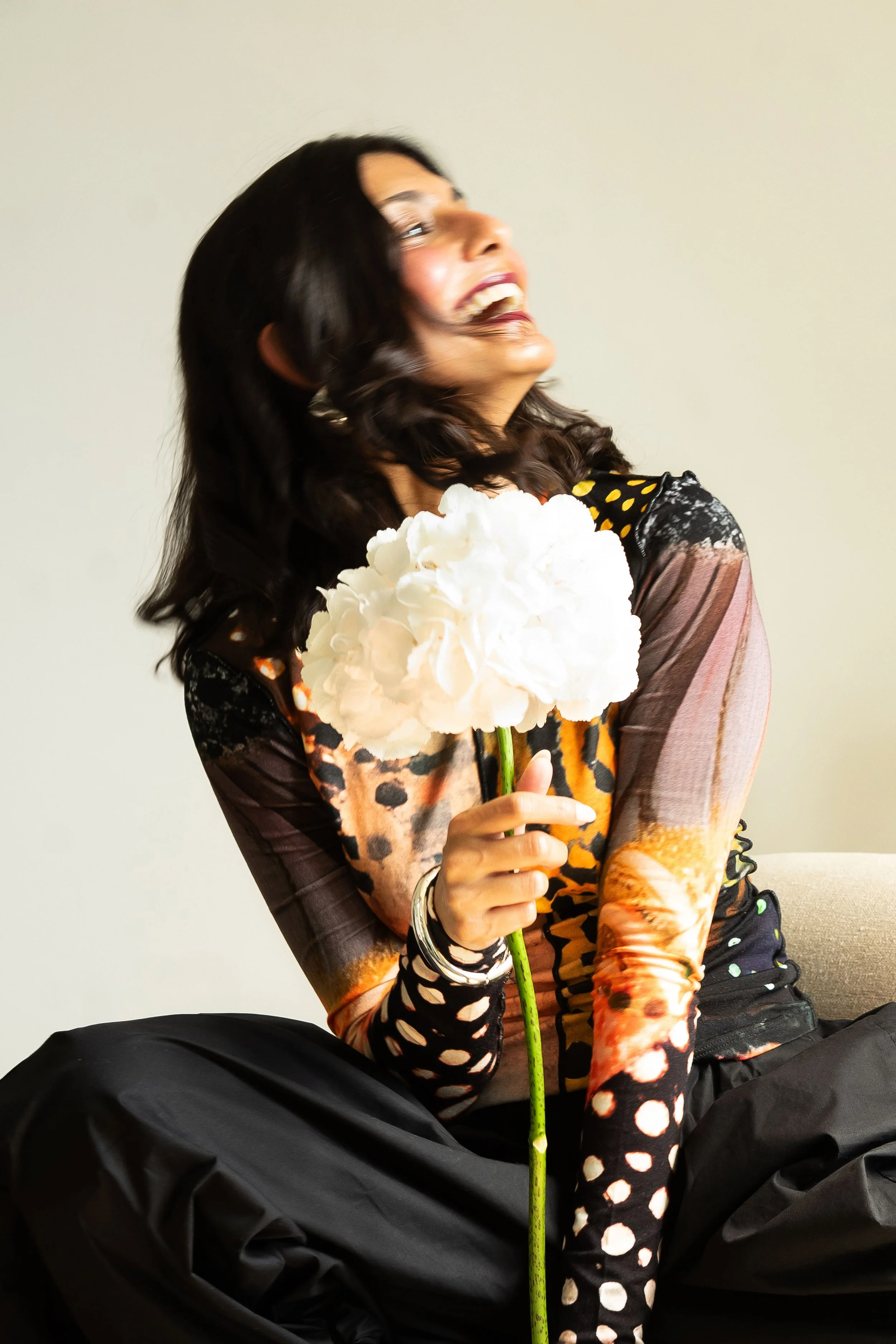 Woman with dark hair smiling while holding a white flower, seated against a plain light background, wearing colorful patterned clothing with polka dots and animal print.