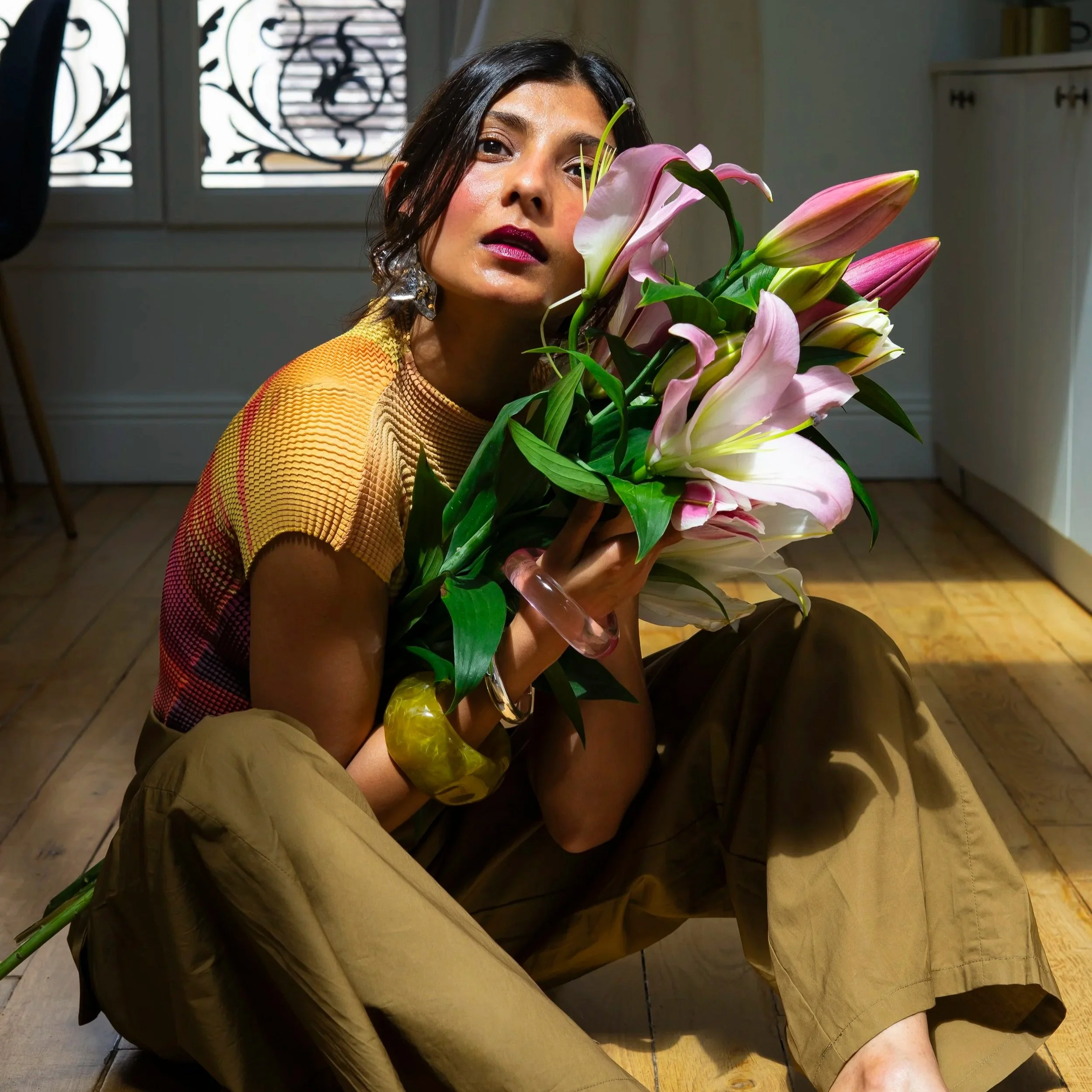 Woman with dark hair holding a bouquet of pink and white lilies, sitting on a wooden floor near a window with decorative ironwork.