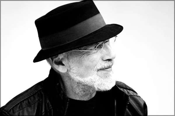 Side profile of an older man wearing glasses, a black hat with a gray band, and a leather jacket.