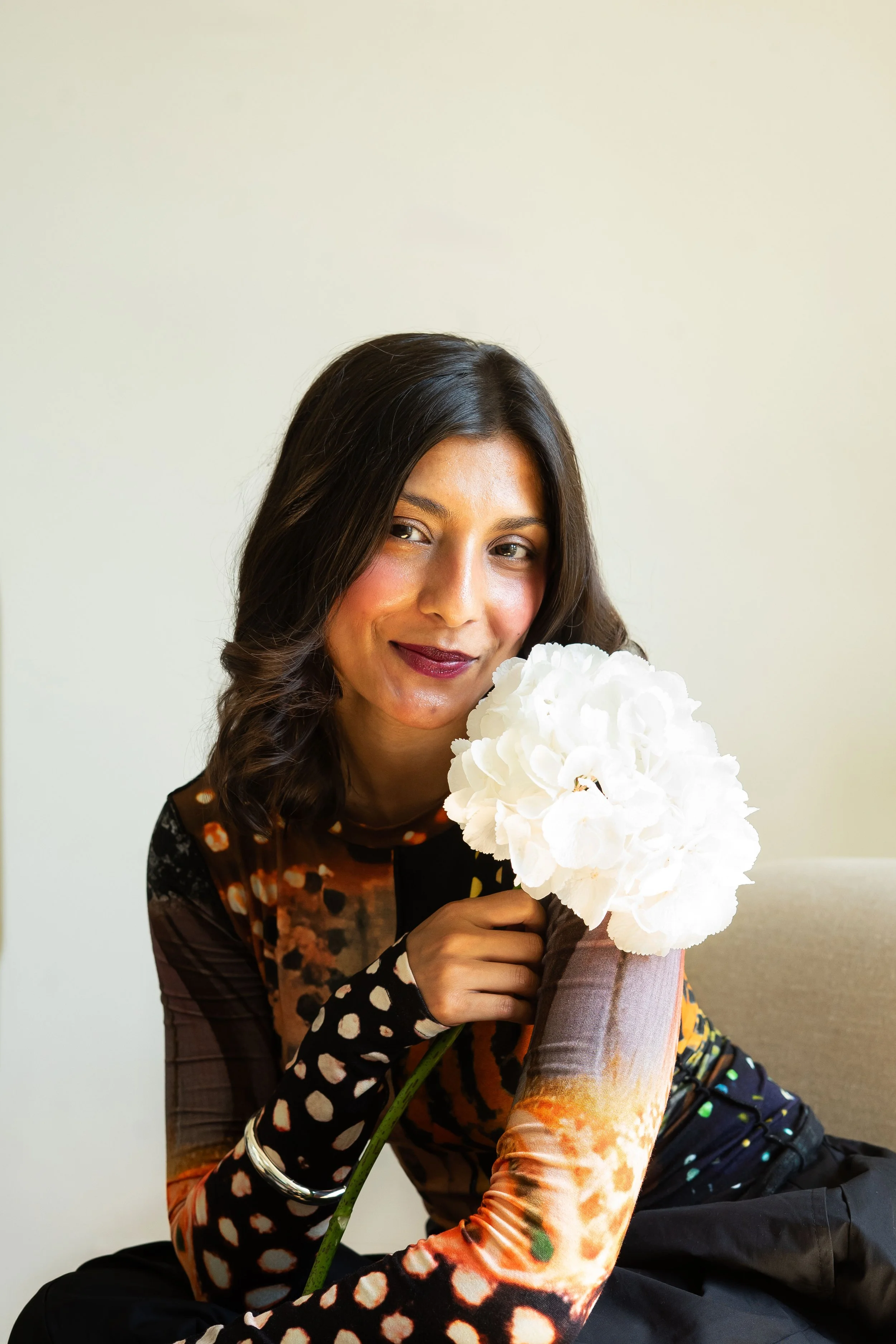 A woman with curly brown hair and a warm smile holding a large white hydrangea flower in front of a plain light-colored background.