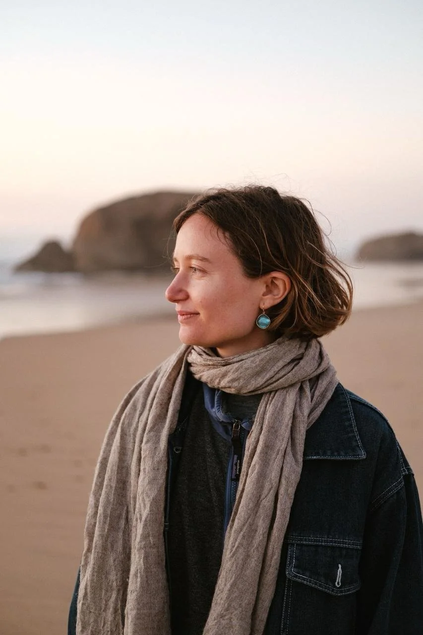 A woman standing on a beach during sunset, wearing a dark jacket, a gray scarf, and blue earrings.