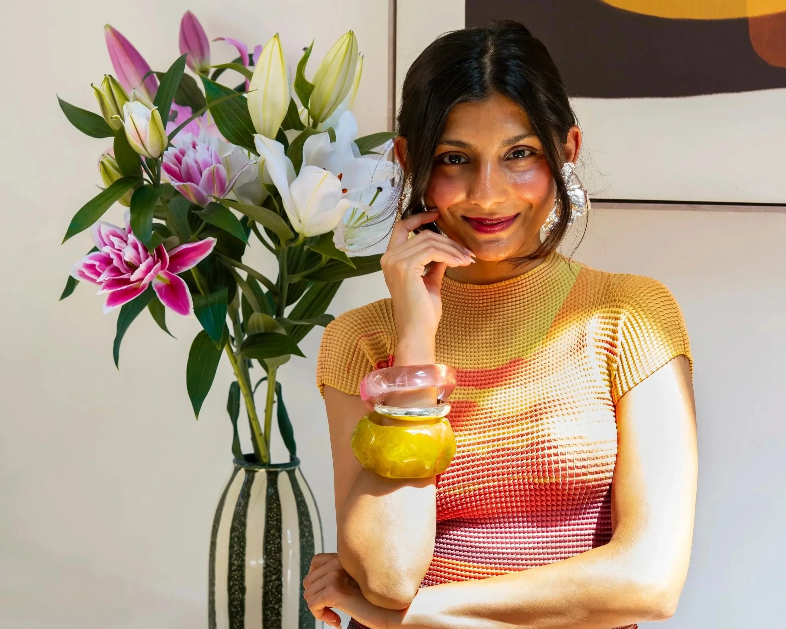 A woman with dark hair, wearing a yellow top, sitting in front of a large bouquet of pink and white lilies, with her chin resting on her hand and smiling.