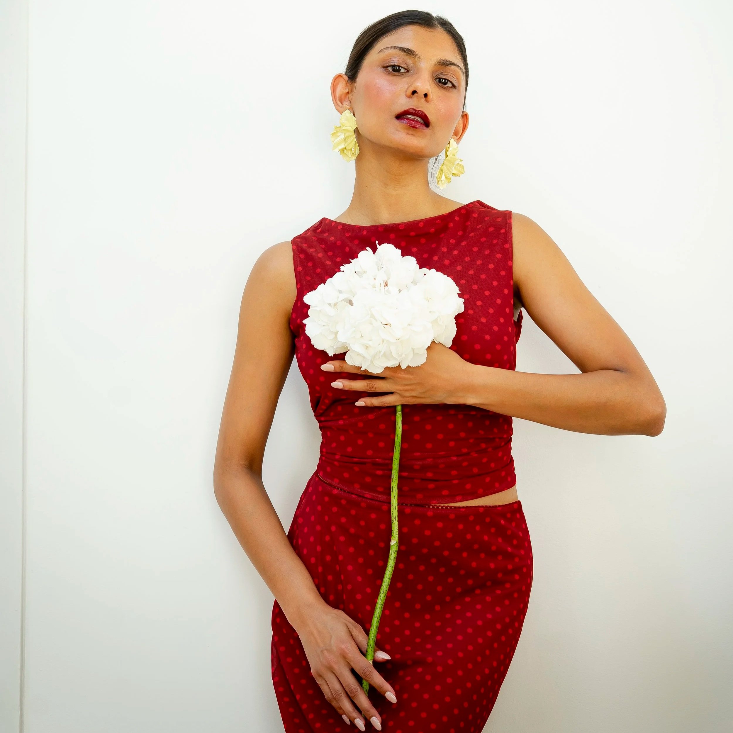 A woman in a red dress with polka dots holds a bouquet of white flowers against a white wall background.
