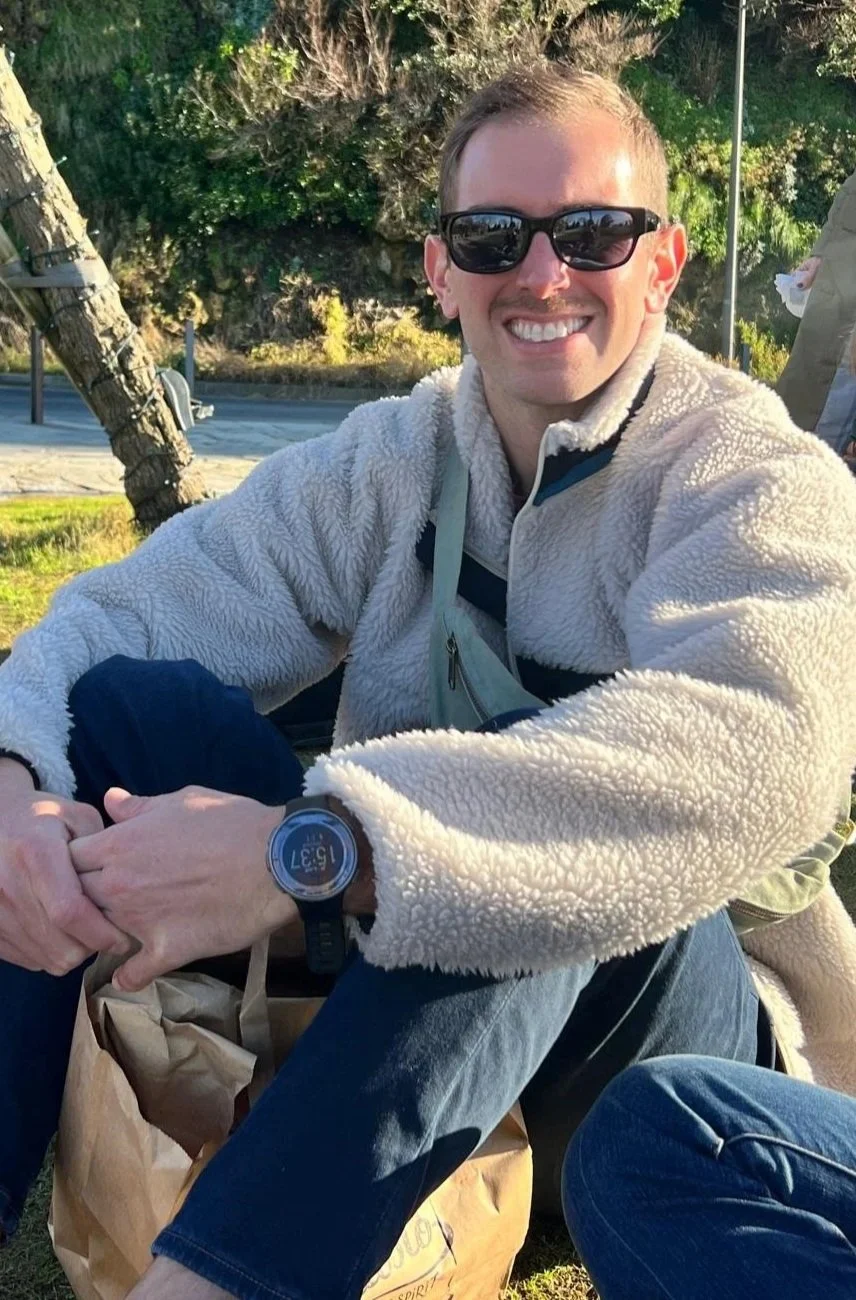 A smiling man wearing sunglasses and a fleece jacket, sitting outdoors in a park-like setting with trees and a bench in the background.