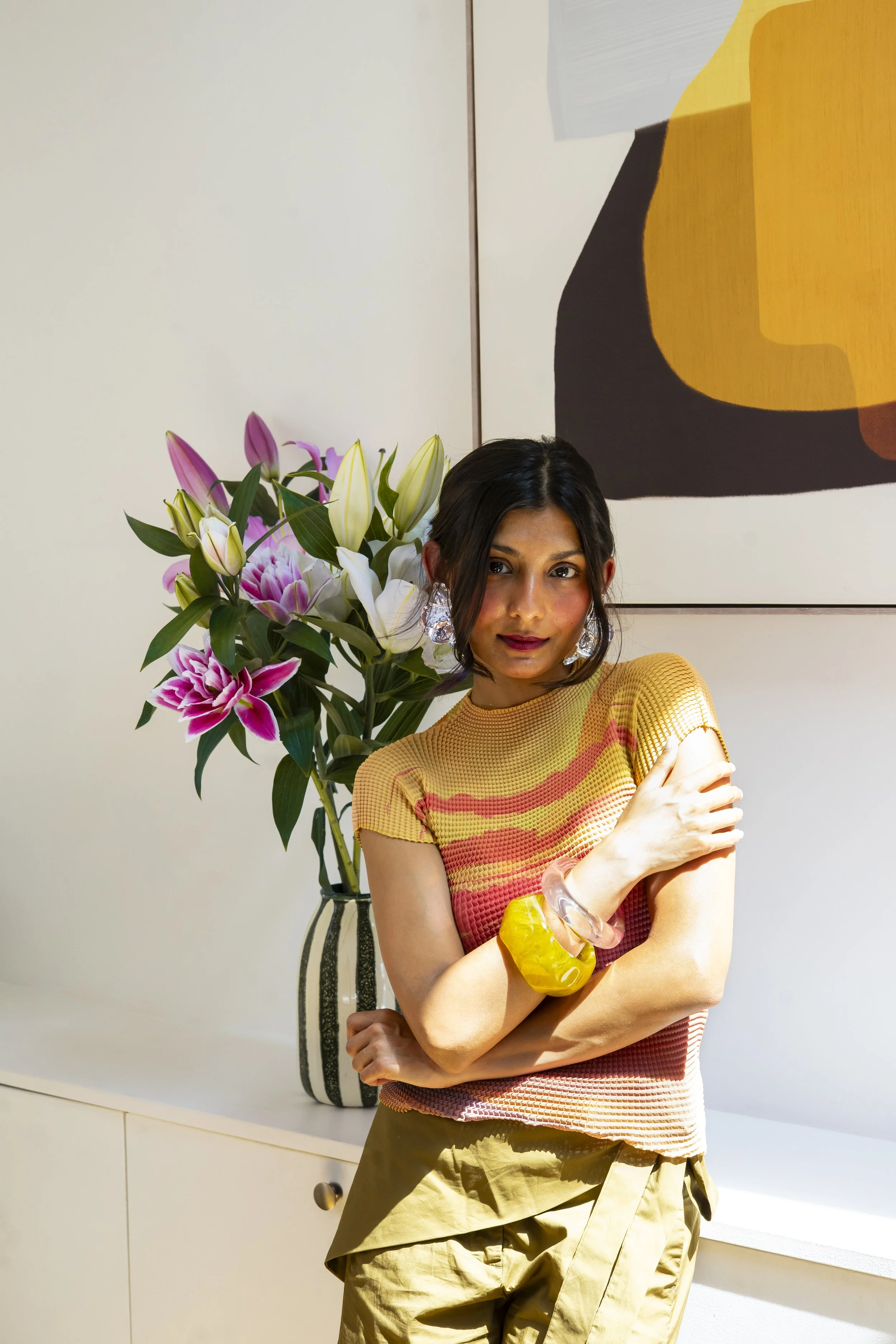 A woman with dark hair wearing a yellow and red checkered top, bright yellow shorts, and clear and yellow bracelets, standing in front of a large bouquet of pink and white lilies with green leaves, in an art-filled indoor space.