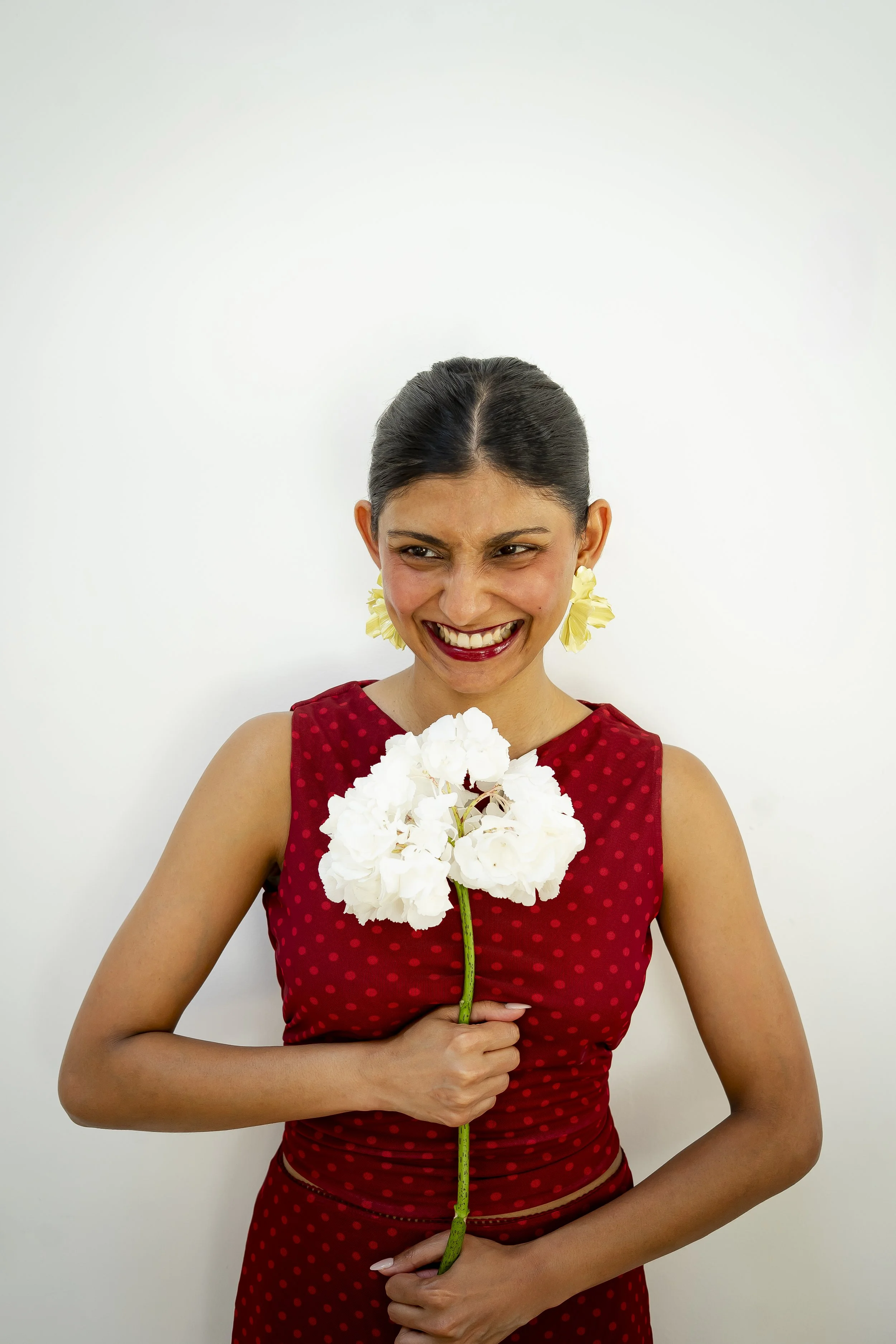 A woman wearing a red dress with small polka dots, holding a white flower close to her chest, smiling and looking down, standing against a plain white background.