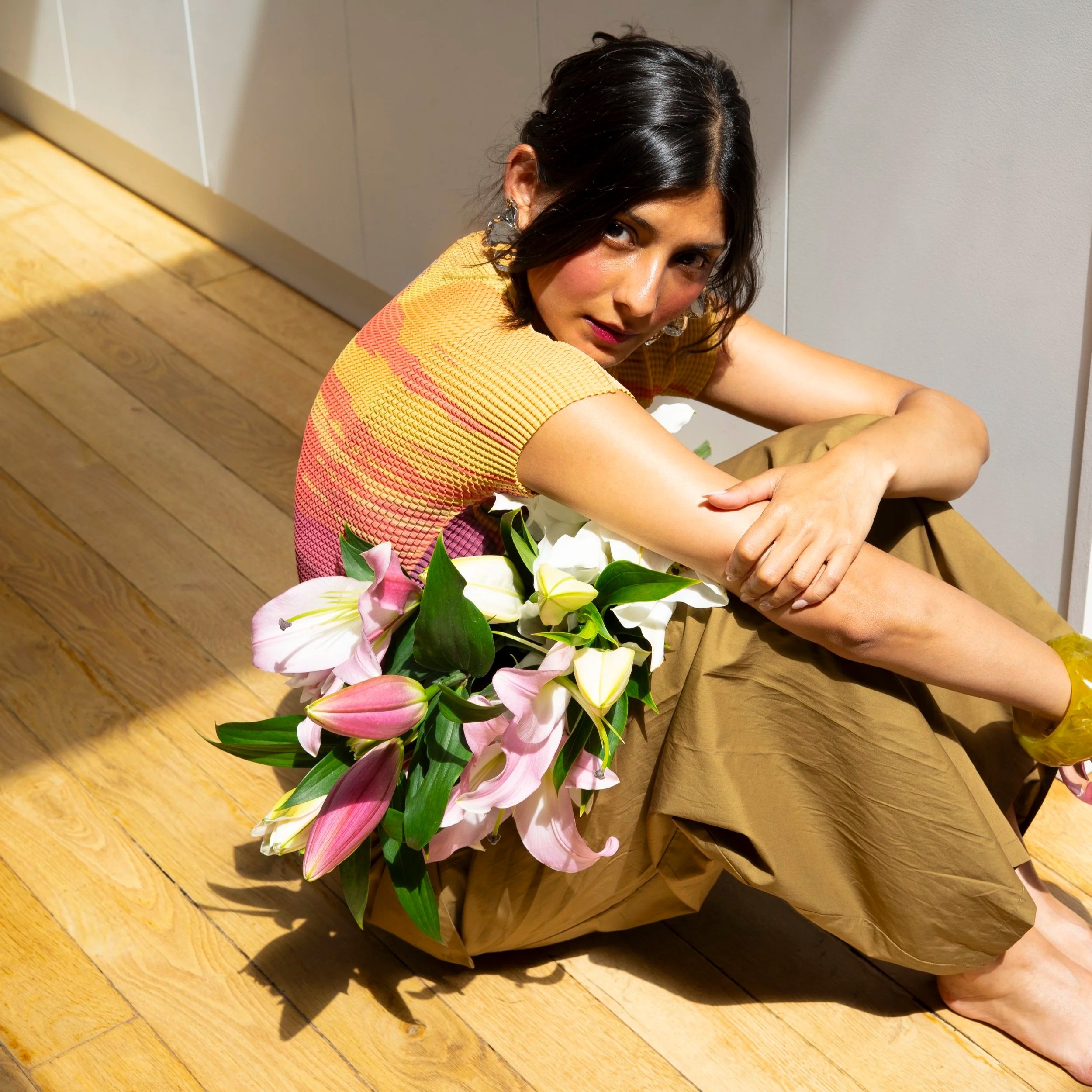 A woman with dark hair and earrings sitting on a wooden floor with a bouquet of pink and white lilies, wearing a yellow and pink patterned top, indoors against a plain wall.
