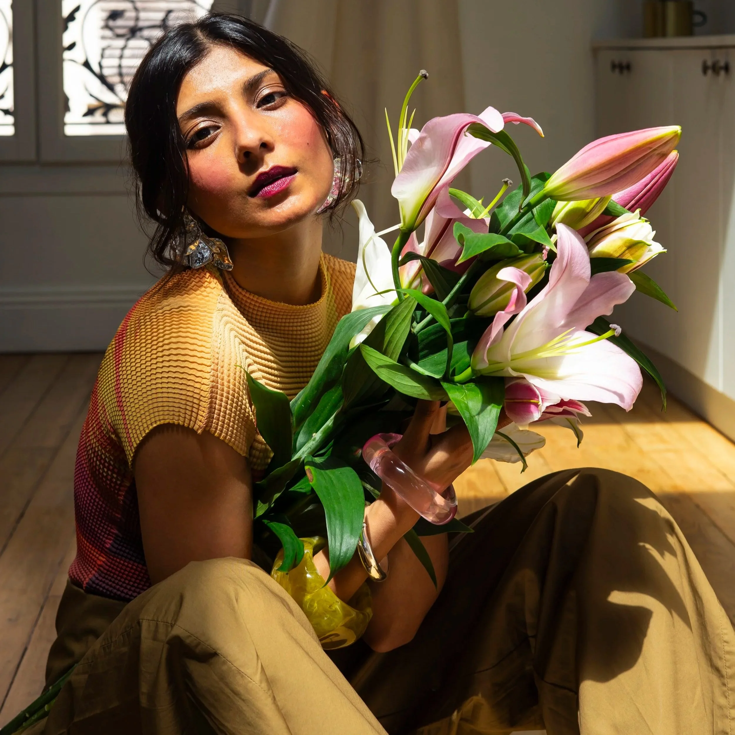 A woman with dark hair and makeup holding a large bouquet of pink lilies and greenery, wearing a yellow textured top and displaying earrings, sitting indoors with a window and furniture in the background.