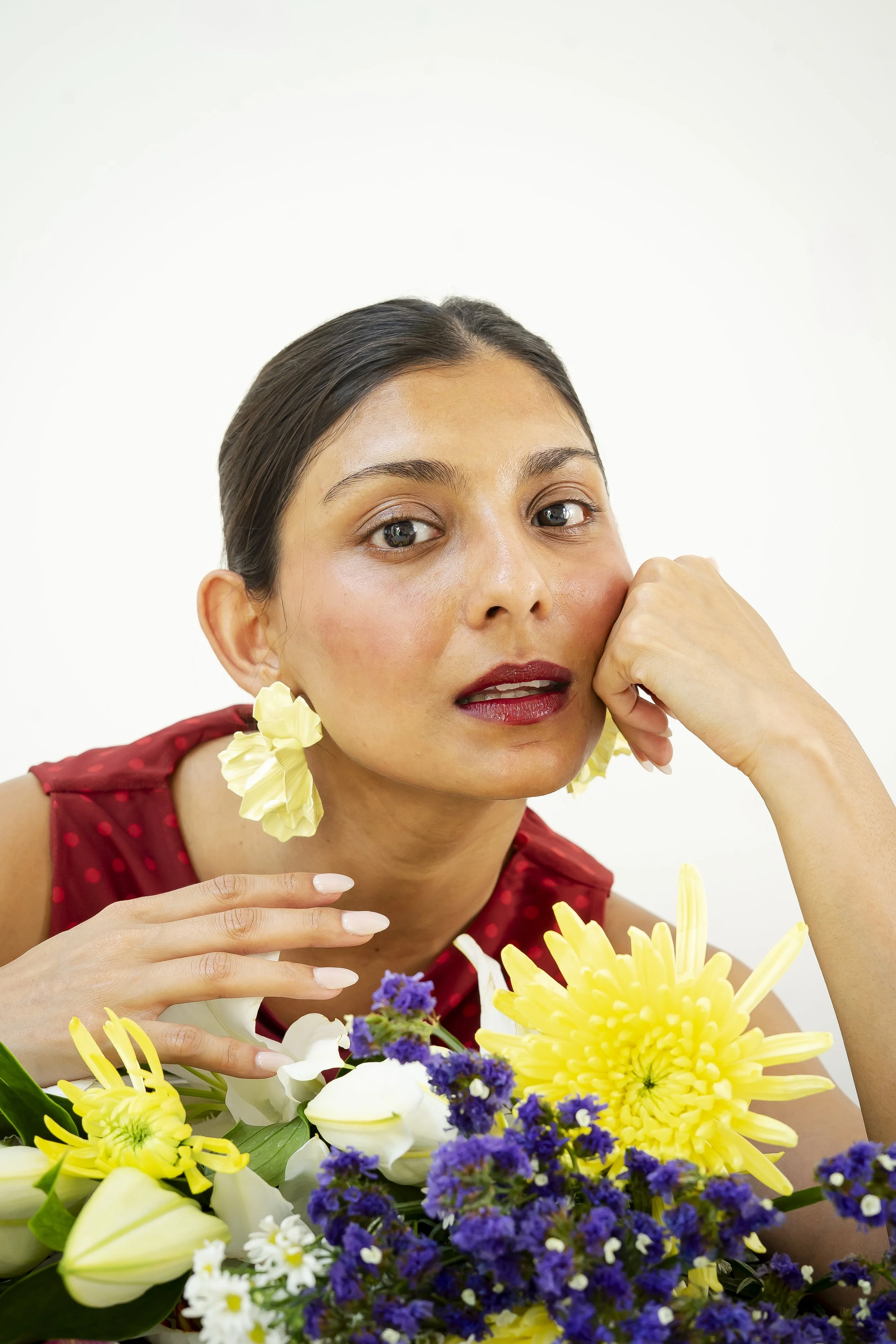 A woman with dark hair and makeup, wearing a red dress with polka dots and large yellow earrings, is posing behind a colorful bouquet of yellow, purple, white, and green flowers with a plain white background.