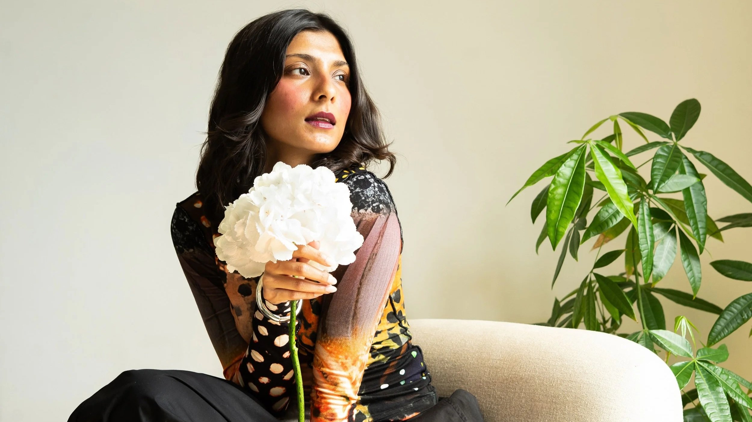 A woman with dark hair sitting on a beige couch, holding a large white flower, with a large green leafy plant beside her, against a neutral background.
