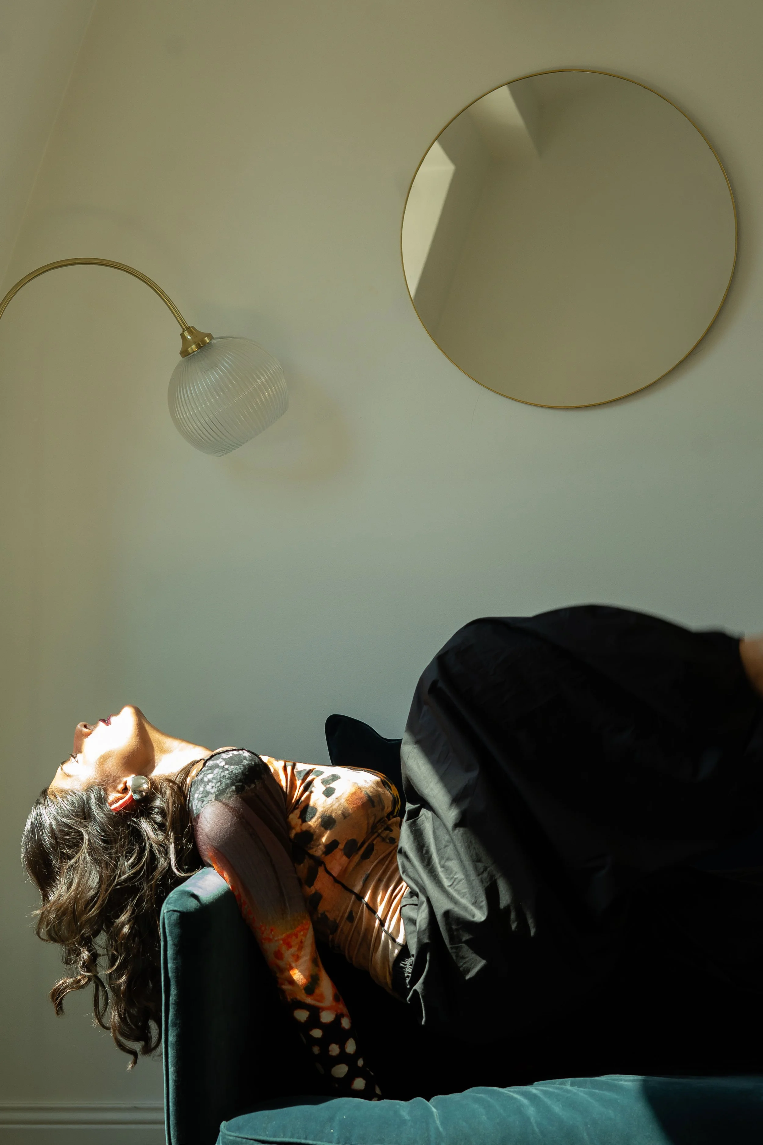 A woman with long, curly hair and large earrings is leaning back on a green armchair with her eyes closed, and her mouth open, laughing or smiling. Her head is tilted back and she appears to be relaxed. Part of a person's legs in dark pants are visible in the foreground, while a circular mirror and a wall-mounted lamp are seen on the wall behind her.