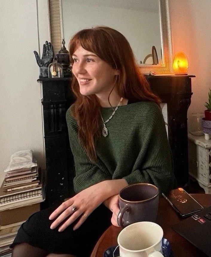 A young woman with long red hair is sitting at a table in a cozy room, smiling and looking to her right. She is wearing a dark green sweater and a silver necklace. On the table are two coffee mugs and a smartphone. The background features a black fireplace mantel with candles, a mirror, and a small lamp, along with some greenery on a side table.
