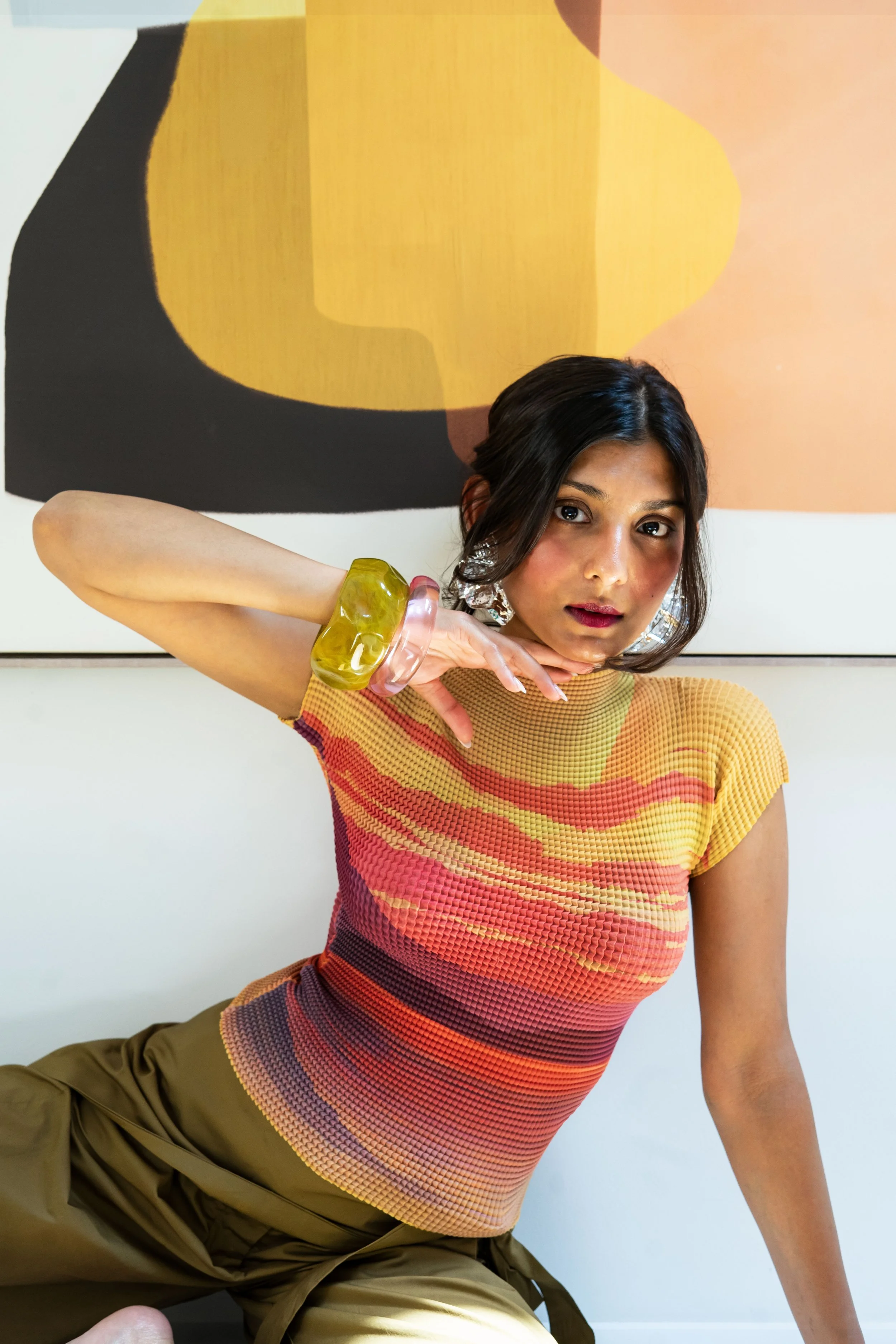 A young woman wearing a multicolored striped top and olive green pants, posing indoors against an abstract yellow and black painting.