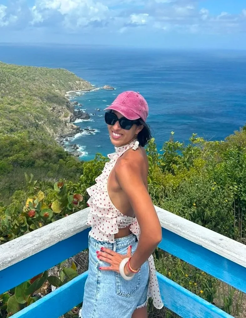 A woman in sunglasses and a pink cap poses on a blue and white railing overlooking the ocean and a green hillside.