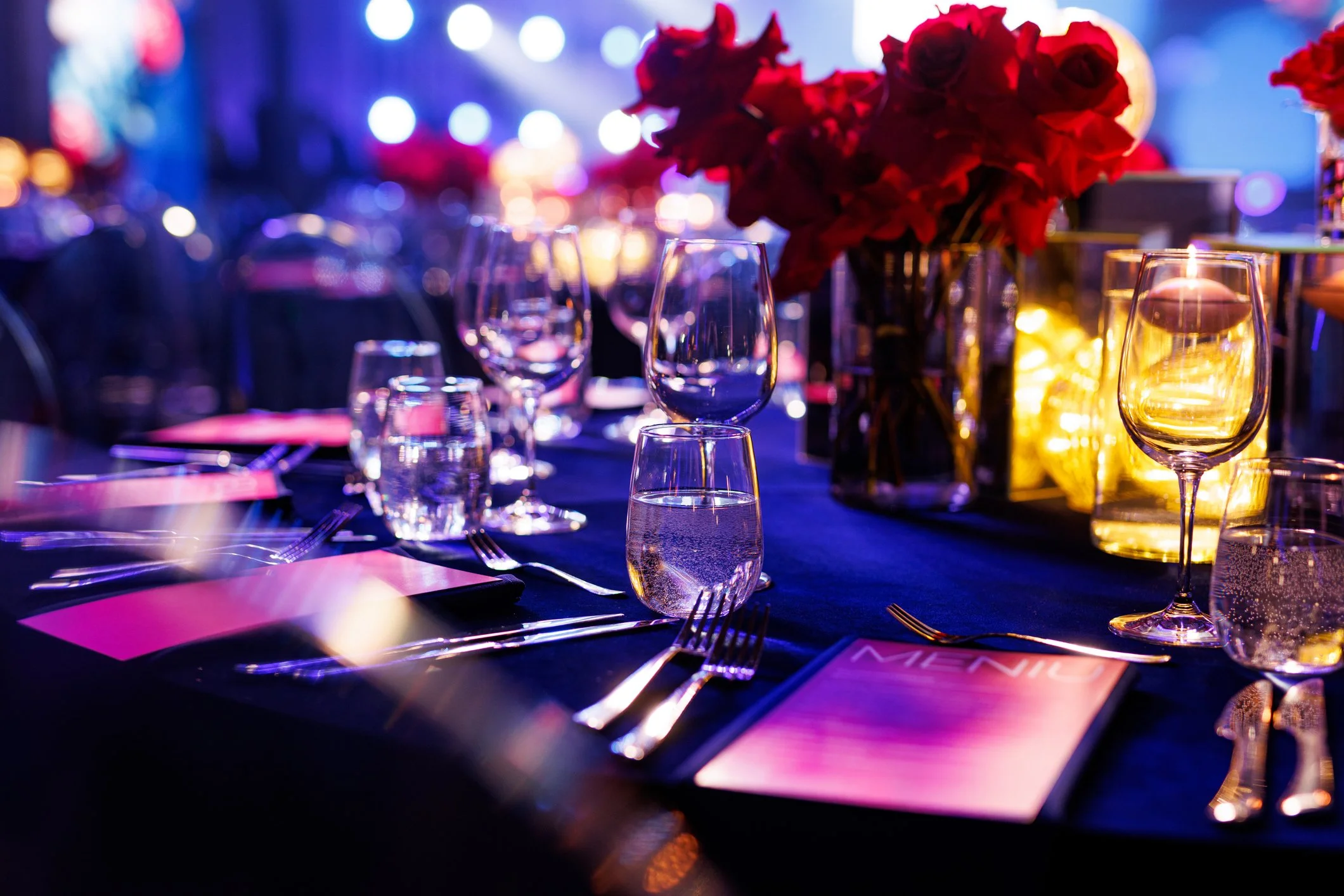 A decorated banquet table set for a formal event with wine glasses, water glasses, silverware, and floral centerpieces with red roses, illuminated by warm and colorful lighting.