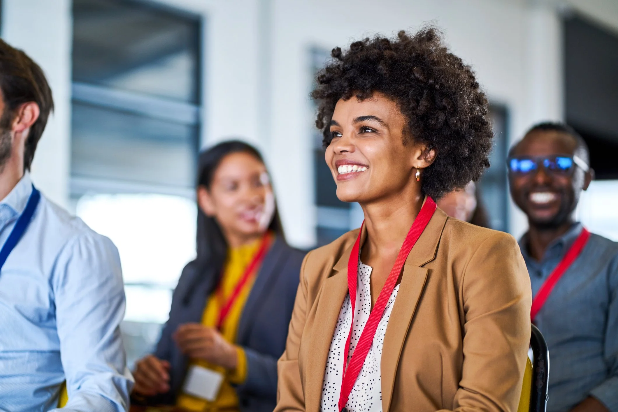 Smiling woman with curly hair wearing a tan blazer participating in a conference or seminar with other attendees.