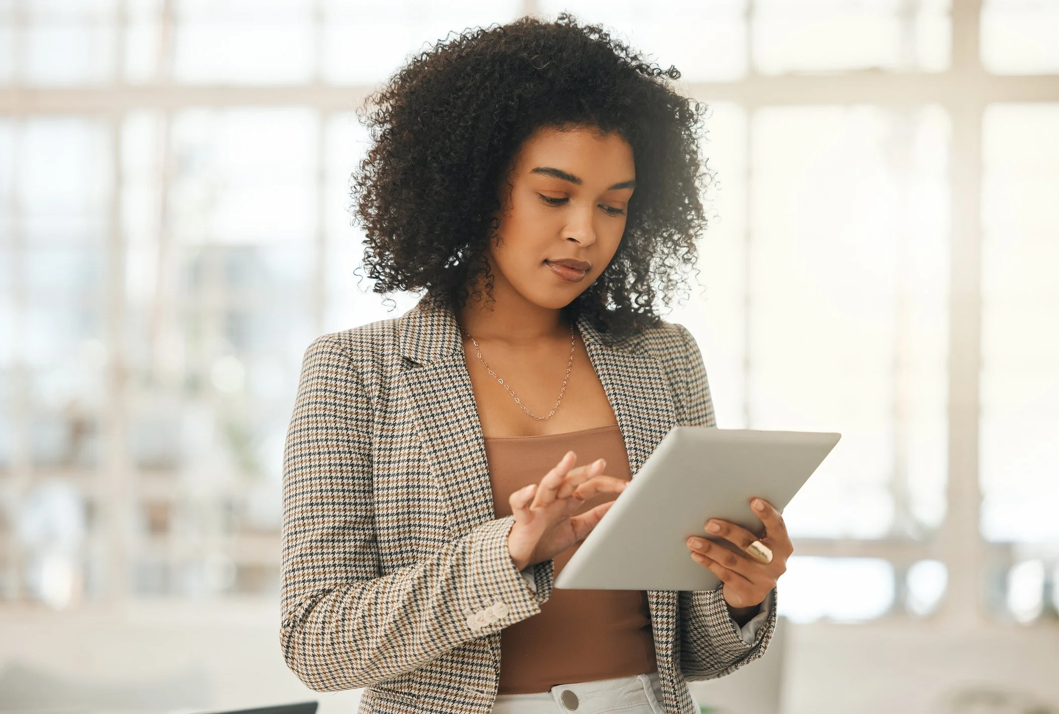 Young woman with curly hair, wearing a checkered blazer and brown top, using a tablet in a bright, modern indoor space.